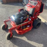 Red Toro GrandStand stand-on mower with engine, controls, and large rear tire on gravel surface, outdoors near a fence gate.