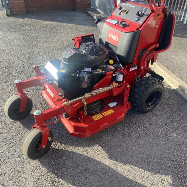 Red Toro GrandStand stand-on mower with engine, controls, and large rear tire on gravel surface, outdoors near a fence gate.