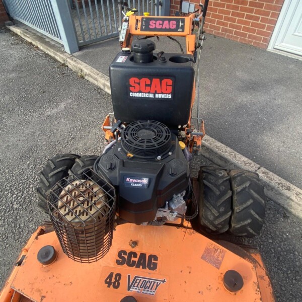Front/center view of an orange SCAG commercial mower with a Kawasaki engine, parked on a paved surface near a brick wall and metal gate, showing a protective grid on the side basket and large rear tires.