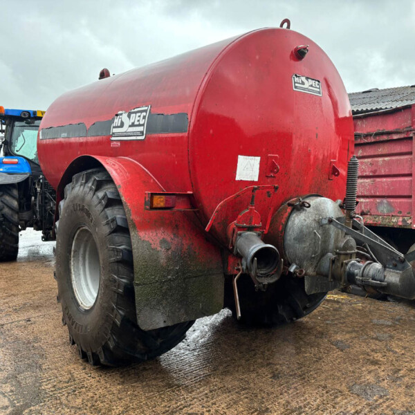 Red cylindrical agricultural tanker attached to a blue tractor in a muddy farm yard.