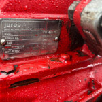 Close-up of a metal identification plate on red machinery, partially wet with water droplets, showing model and serial information.