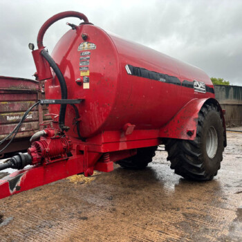 Red agricultural sprayer/tanker on a trailer with hoses and gauges in a muddy lot, under a cloudy sky.