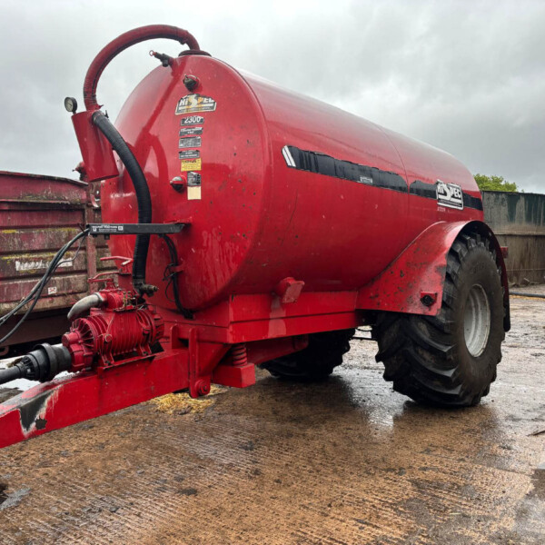 Red agricultural sprayer/tanker on a trailer with hoses and gauges in a muddy lot, under a cloudy sky.