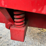 Close-up of red machinery showing a coiled metal spring and angled plates, outdoors on gravel ground.