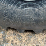 Close-up of a worn tire sidewall with raised alphanumeric markings, sitting on muddy ground.