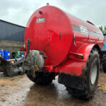 Red cylindrical tanker trailer hitched to a blue tractor in a muddy industrial yard.
