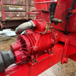 Close-up of a red hydraulic pump and drive shaft on heavy farm equipment, with wet grime and mud on the components and ground nearby.