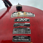 Close-up of a red industrial tank dome with worn warning labels and the number plate '2300' visible, viewed from above against a cloudy sky.