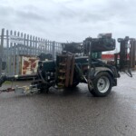Trailer-mounted industrial machine with hoses and metal framework in a fenced storage yard behind a tall metal picket fence.