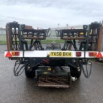 Back of a flatbed trailer carrying rusty farm equipment with hoses and metal frames, license plate YX58 BKN, in an industrial yard with a fence behind.