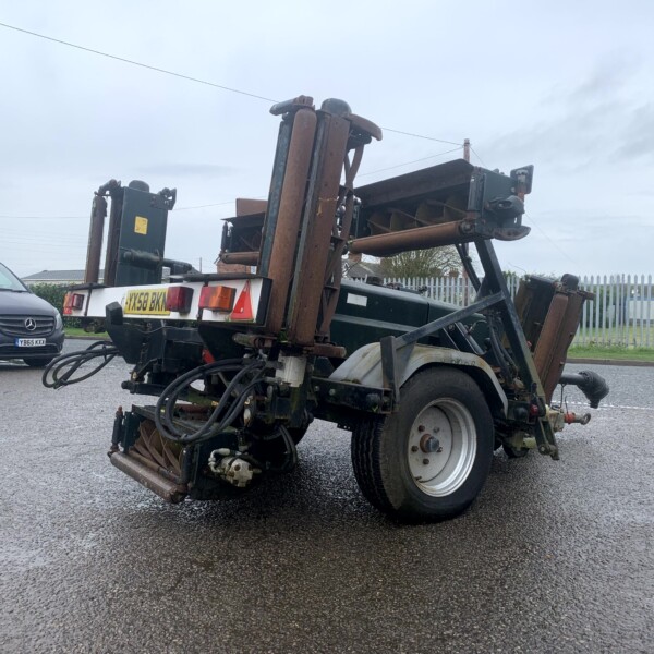 Rusty industrial trailer with hydraulic arms and hoses in a parking lot beside a gray van on a wet surface.