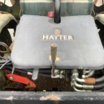 Close-up of a dusty Hayter lawn tractor engine cover with exposed hoses and a red battery jumper clamp nearby.
