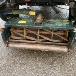 Rusty green agricultural implement attached under a trailer, with rusted hitch, warning labels, and weathered metal parts on a gravel surface.