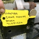Close-up of a yellow inventory tag on a rusty mower part, with handwritten words and a coin tucked in the edge for scale.