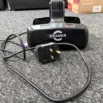 Black Cramer charging base on a gray carpet with a power cord and plug in the foreground and cardboard boxes in the background