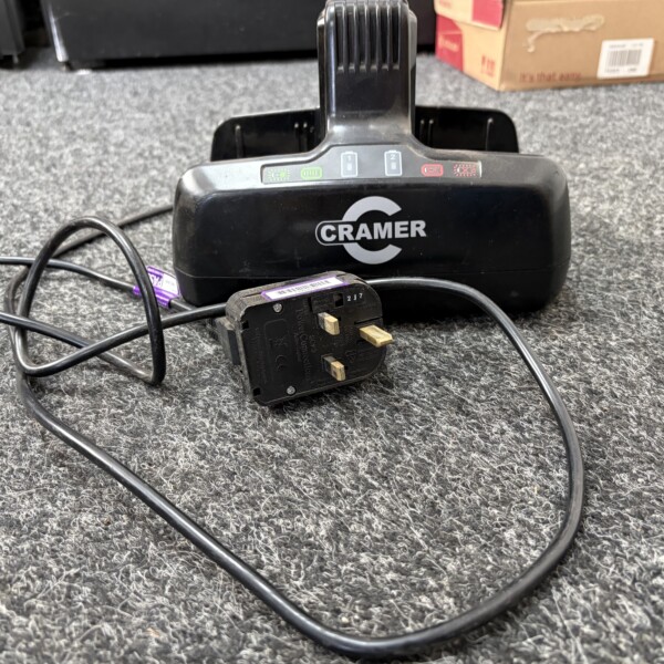 Black Cramer charging base on a gray carpet with a power cord and plug in the foreground and cardboard boxes in the background