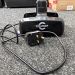 Black CRAMER charging base on a gray carpet with tangled power cords and a plug-in adapter nearby.