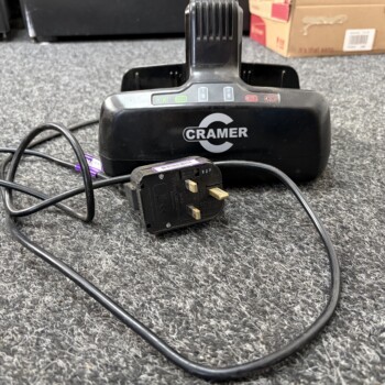Black CRAMER charging base on a gray carpet with tangled power cords and a plug-in adapter nearby.