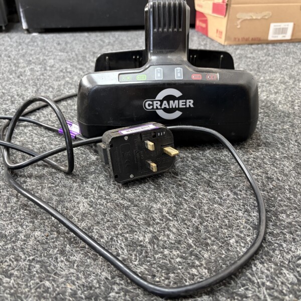 Black CRAMER charging base on a gray carpet with tangled power cords and a plug-in adapter nearby.