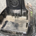 Close-up of a dusty black handheld power tool showing a circular vent/grill and a row of metal prongs at the base, with green indicator icons on the side.