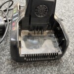 Base charging cradle with plug prongs and a dusty black plastic housing on a gray carpeted floor, vent at back panel.