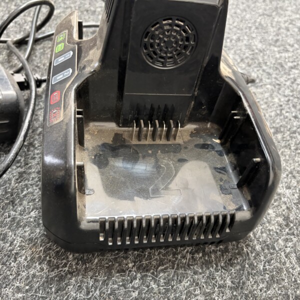 Base charging cradle with plug prongs and a dusty black plastic housing on a gray carpeted floor, vent at back panel.