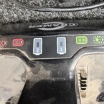 Close-up of a car dashboard control panel with red warning icons on the left and a green battery indicator on the right; a black cable runs across the top.