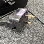 Close-up of a black battery charger with a purple barcode label and brass prongs on a speckled gray surface.