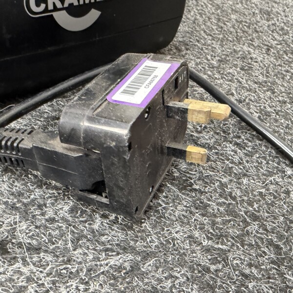 Close-up of a black battery charger with a purple barcode label and brass prongs on a speckled gray surface.