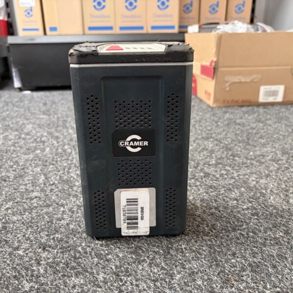 Close-up of a black, worn electronic device with perforated vents and a CRAMER logo, barcode label visible, standing on a gray carpet in a store aisle.