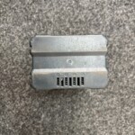 Close-up of a dusty gray electronic module with vent slots and a single screw on a dark carpeted floor