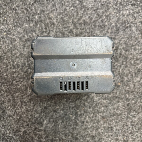 Close-up of a dusty gray electronic module with vent slots and a single screw on a dark carpeted floor
