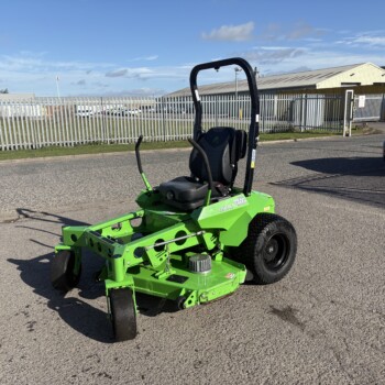 Bright green riding lawn mower parked on a street with a metal fence and industrial buildings in the background under a blue sky.