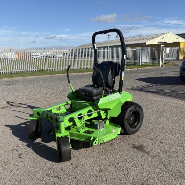 Green ride-on mower in the foreground on a paved lot, with a tall metal fence and industrial warehouses in the background under a blue sky.