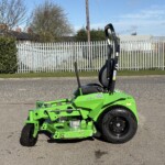 Bright green riding lawn mower on a street, with a metal fence and blue sky in the background.