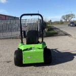 Rear view of a bright green riding lawn mower with a black seat on a paved area, facing a tall metal fence and a building with signboards in a sunny outdoor setting