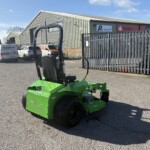 Green riding lawn mower in a parking lot outside a metal industrial warehouse.