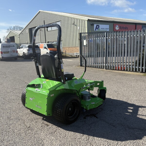 Green riding lawn mower in a parking lot outside a metal industrial warehouse.