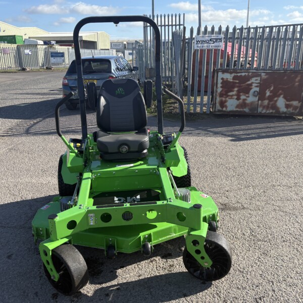 Front view of a bright green ride-on lawn mower in an industrial yard with metal fencing and a parked car in the background, under blue sky.