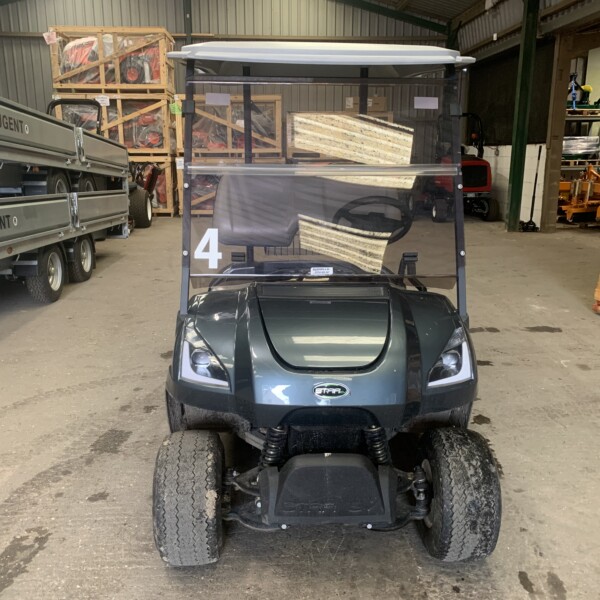 Electric golf cart with a clear canopy positioned in a warehouse, facing the camera, with pallets and machinery in the background.