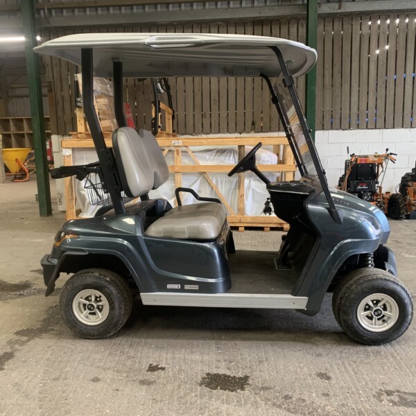 Green golf cart with beige seats and roof, parked inside a warehouse with pallets and orange machinery in the background, side view