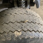 Close-up of a dirty vehicle tire showing the zigzag tread pattern and dirt in the tread. Behind it, part of the suspension coil is visible.