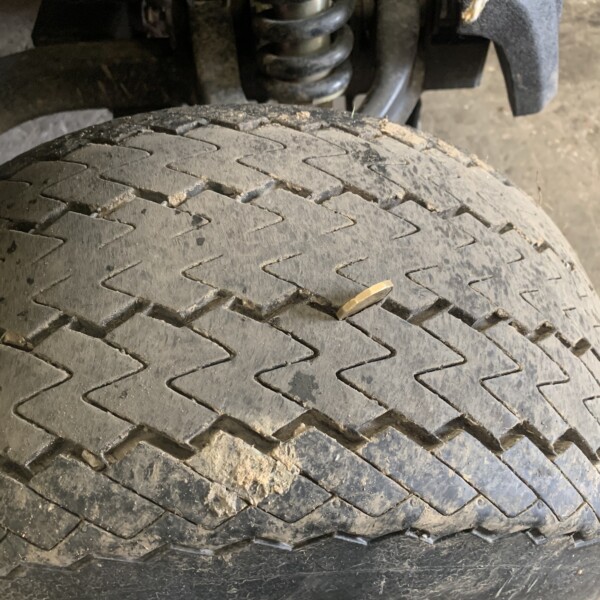 Close-up of a dirty vehicle tire showing the zigzag tread pattern and dirt in the tread. Behind it, part of the suspension coil is visible.