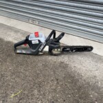 Cordless chainsaw lying on pavement in front of a gray metal roller shutter.