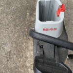 Close-up of a gray 82V cordless power tool base with a red trigger, resting on a black stand on concrete ground.