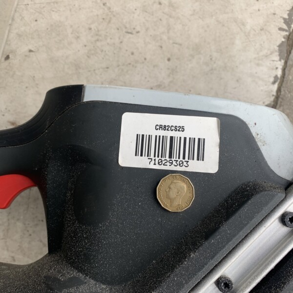 Close-up of a gray and black power tool with a barcode label, a red trigger, and a coin resting on its surface in a dusty workshop area