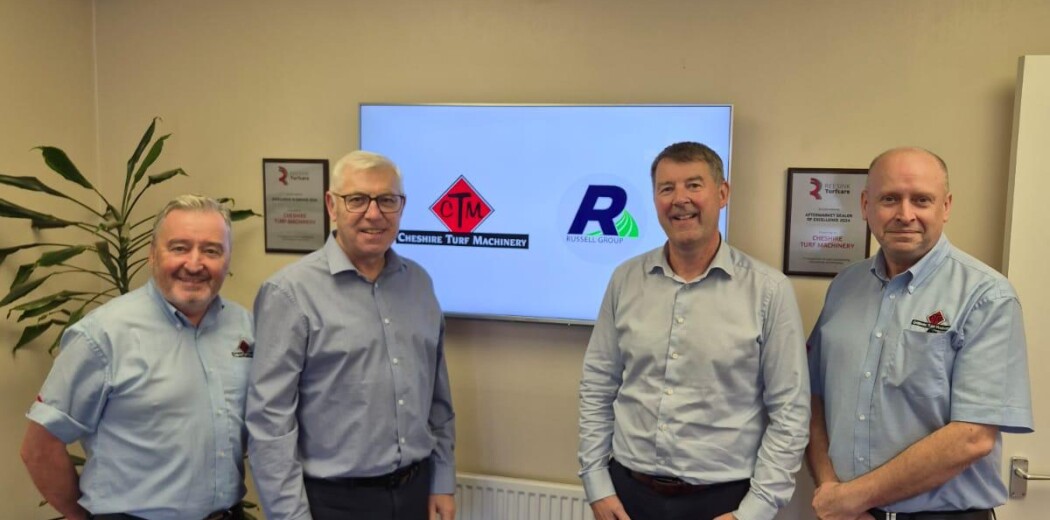 Four men in light blue shirts pose in an office, standing in front of a wall-mounted screen displaying company logos.