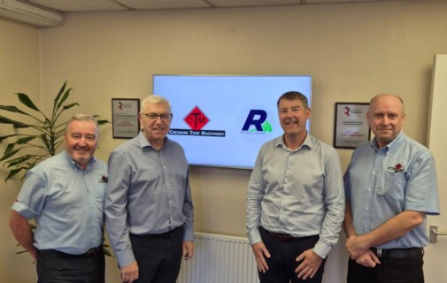 Four men in light blue shirts pose in an office, standing in front of a wall-mounted screen displaying company logos.