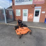 Orange riding lawn mower parked on a paved area outside a brick building with 'RUSSELLS' and 'TORO' signs above the entrance and a reception door nearby