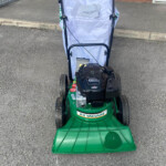 Green KV Vacuum lawn mower with a white bagging grass catcher attached, on a driveway in front of a brick building.